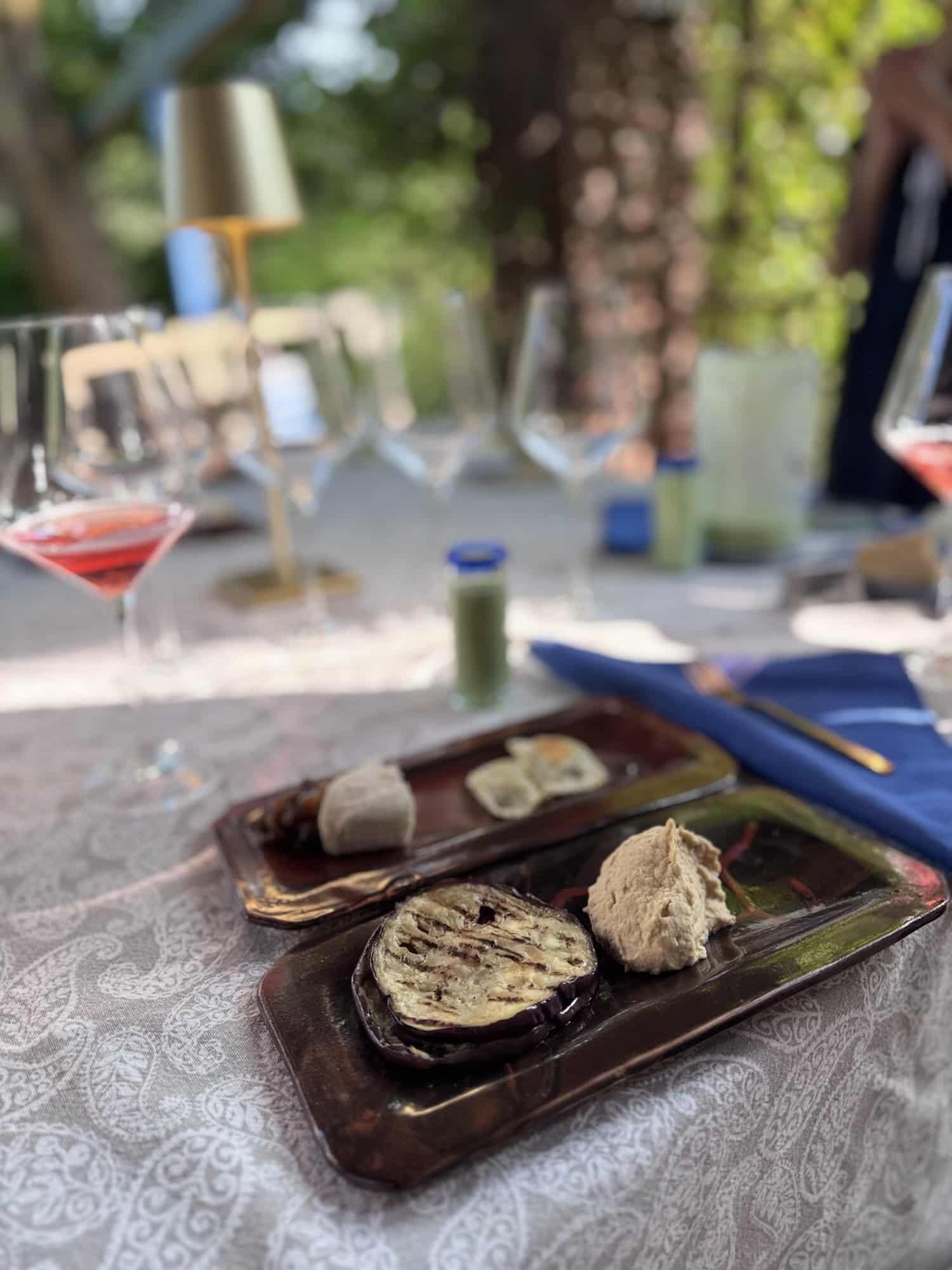 A beautifully arranged plate with grilled eggplant and various dips, set on a textured tablecloth, with wine glasses and a decorative lamp in the background, showcasing a tranquil dining experience outdoors.