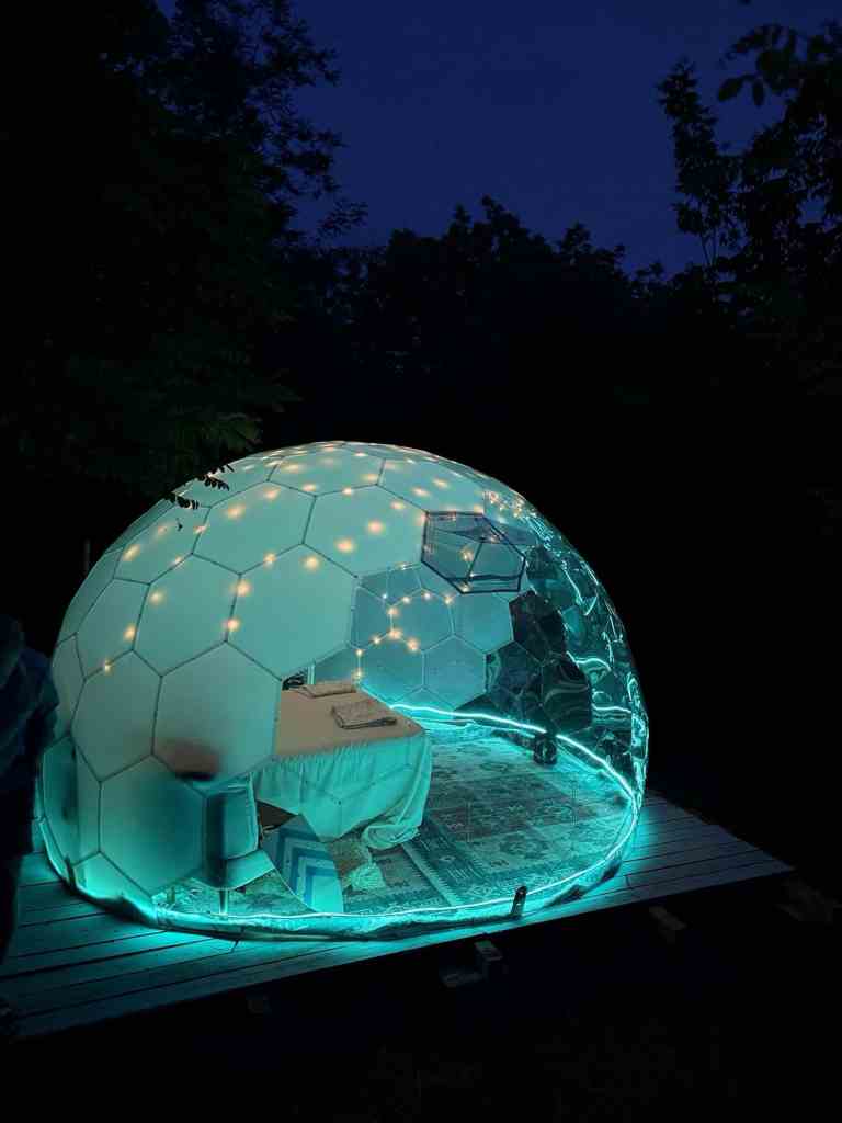 A uniquely designed transparent dome illuminated with soft lights, featuring a cozy interior setup with a table and chairs, surrounded by greenery in a twilight setting.